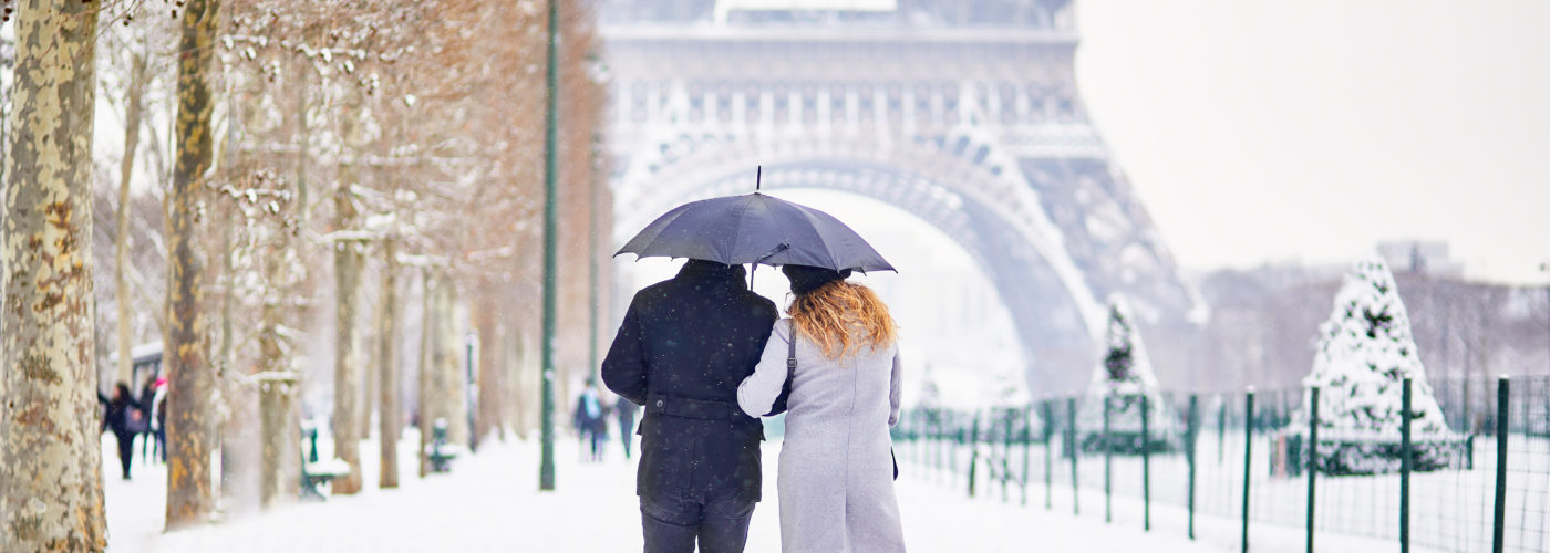 Couple walking along a tree-lined path toward the Eiffel Tower in the snow