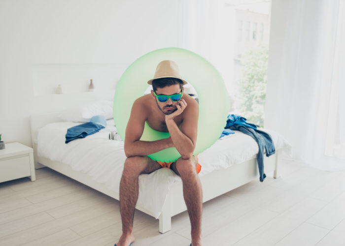 Man wearing swimsuit and inner tube sitting on a hotel bed, looking bored and upset