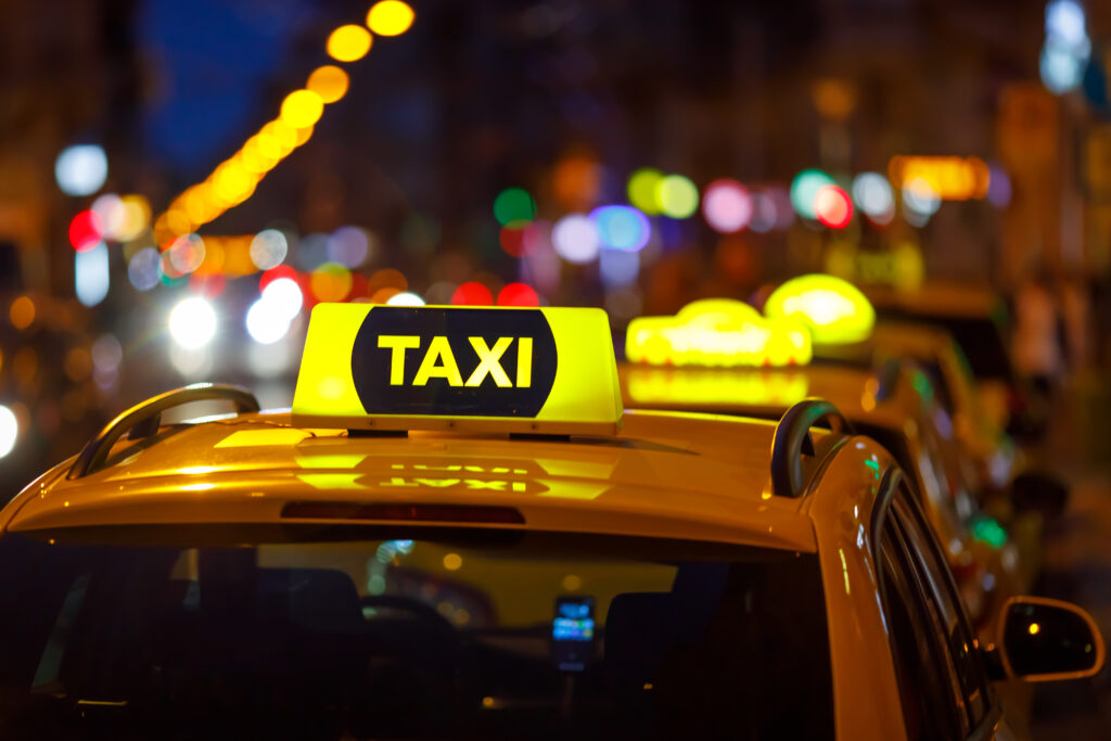 Close up on illuminated yellow taxi sign on top of car with colorful lights blurry in the background