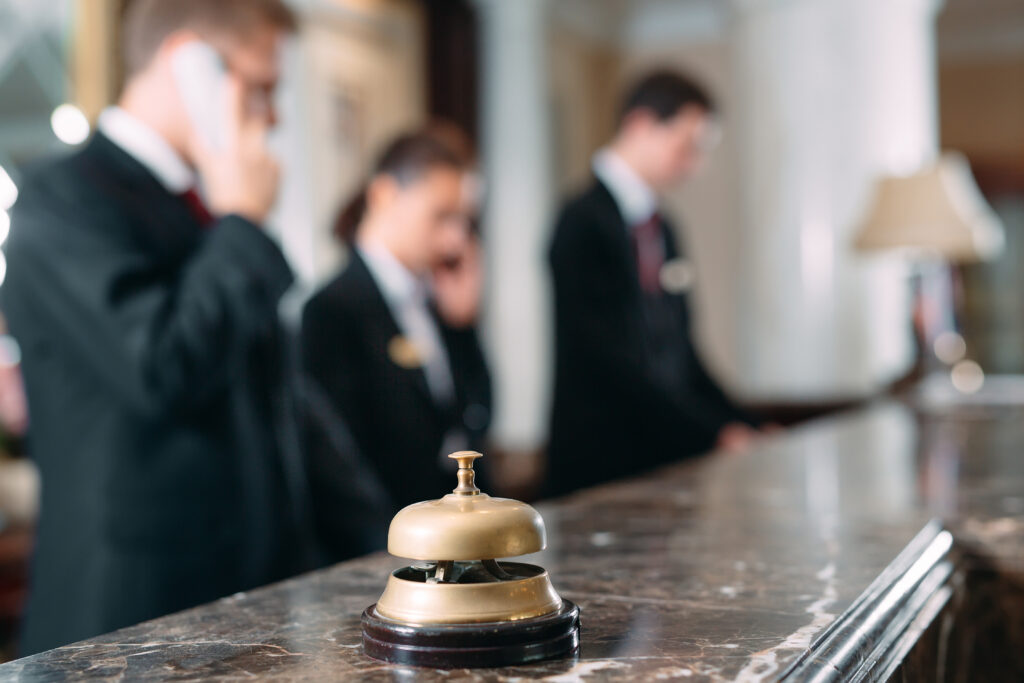 A hotel desk bell in focus in the foreground with hotel staff at the front desk out of focus in the background